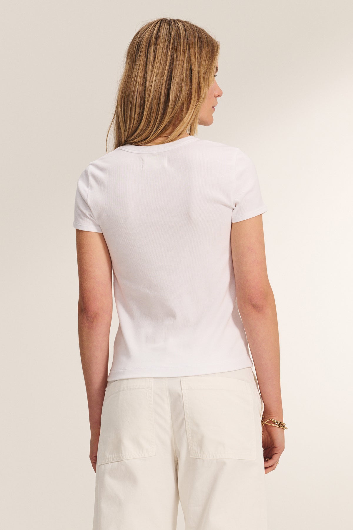 A woman with light brown hair faces away, wearing the BEDFORD TEE by Velvet by Jenny Graham—a plain white short-sleeve modal cotton blend shirt—paired with off-white pants against a neutral background.-39172744052929