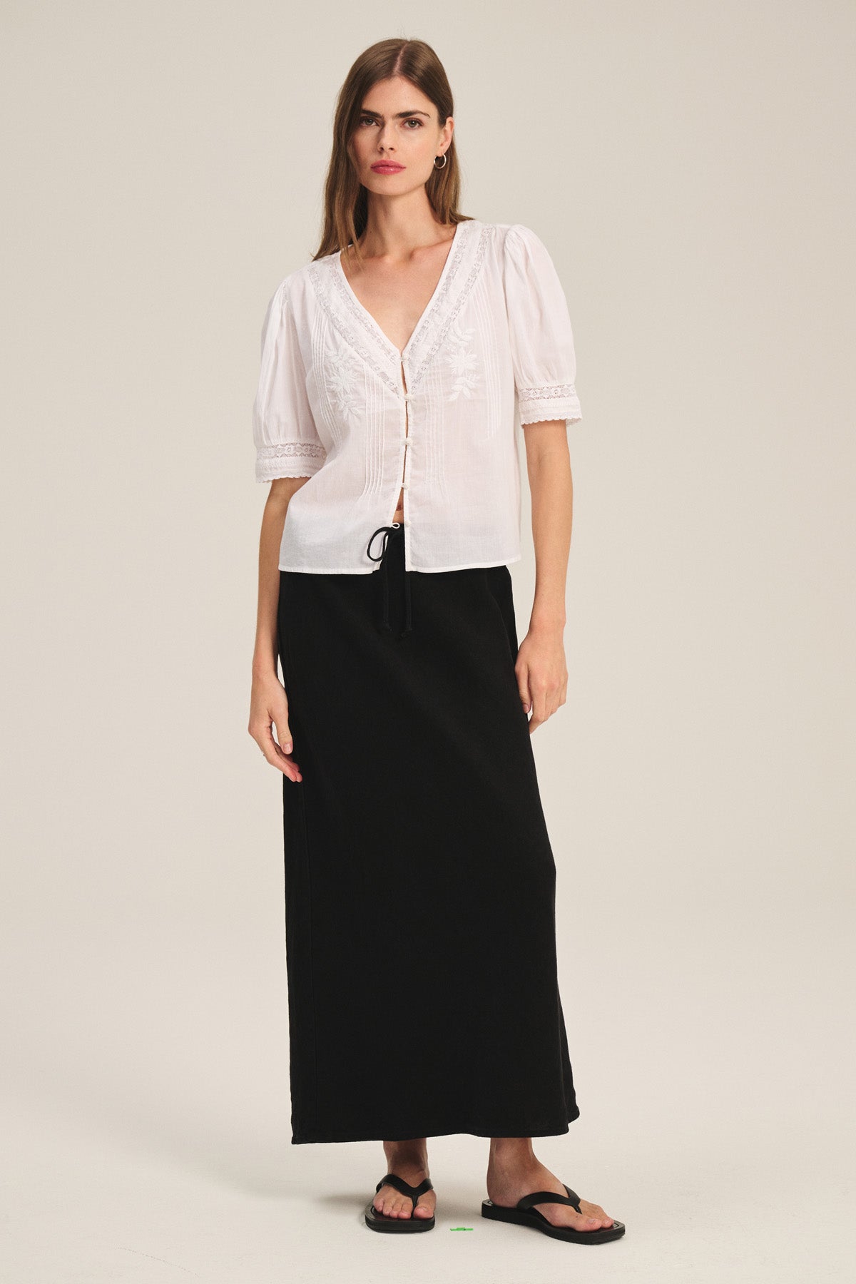 A woman stands against a plain background wearing a white embroidered short-sleeve blouse, black sandals, and the BEKA HEAVY LINEN SKIRT by Velvet by Graham & Spencer—a long black heavy linen maxi skirt with a drawstring elastic waist.-41389447807169