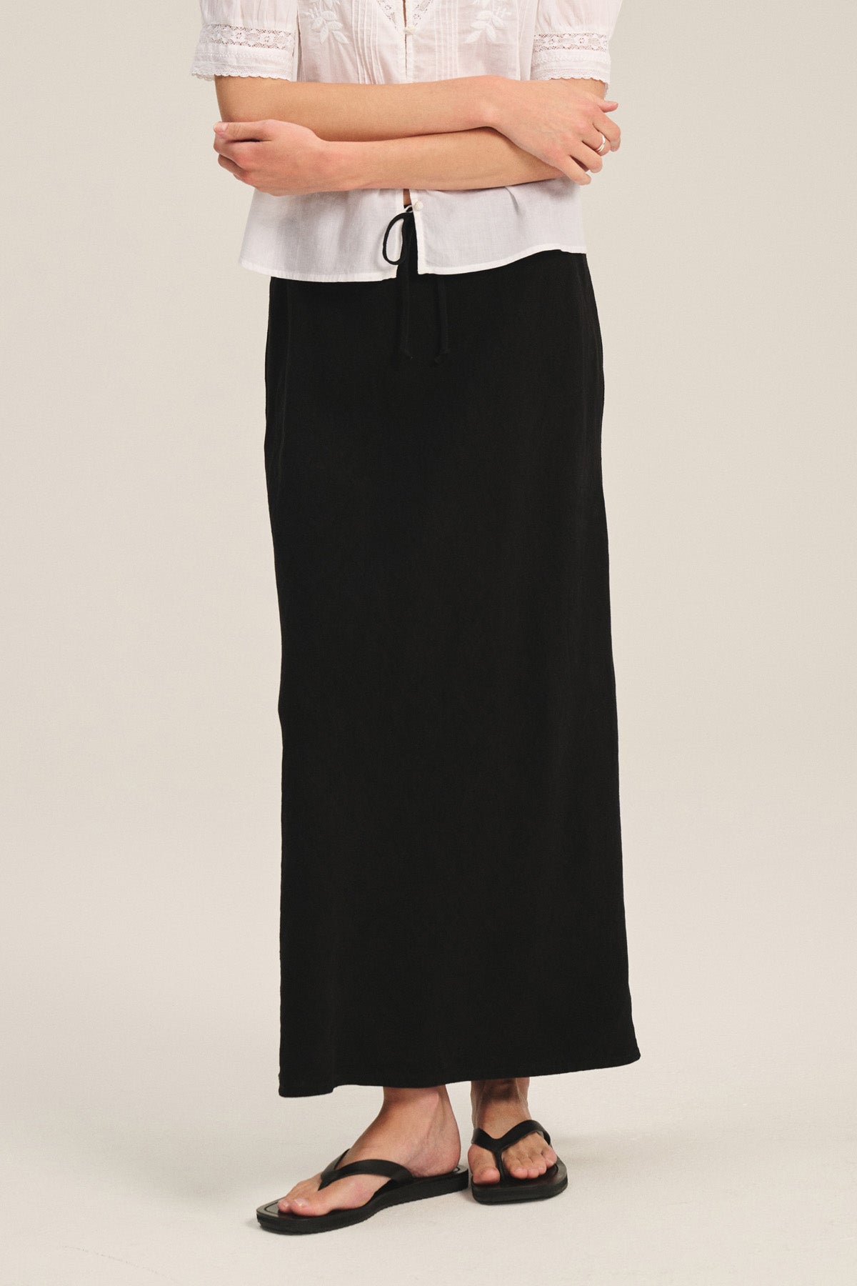 A person wearing a white short-sleeve blouse, the BEKA HEAVY LINEN SKIRT by Velvet by Graham & Spencer with a drawstring elastic waist, and black sandals stands with arms crossed.-41389447774401