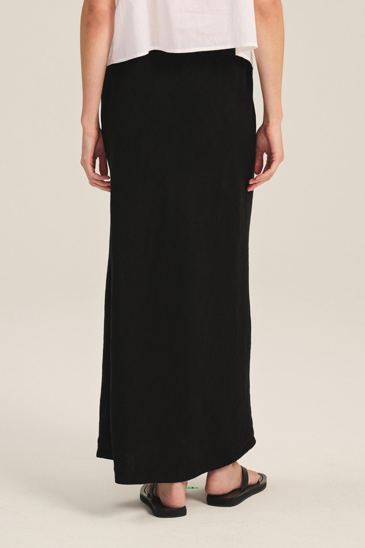 A person wearing a white top, black sandals, and the BEKA HEAVY LINEN SKIRT by Velvet by Graham & Spencer, with its long black heavy linen fabric and drawstring elastic waist, stands facing away from the camera.-41389455835329