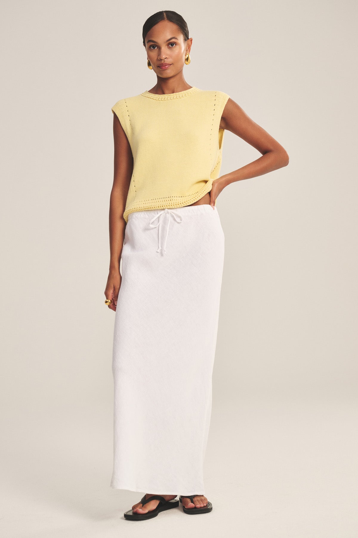 A woman wears the BEKA HEAVY LINEN SKIRT by Velvet by Graham & Spencer—a long white maxi skirt with a drawstring waist—paired with a sleeveless yellow knit top, black sandals, and gold hoop earrings.
