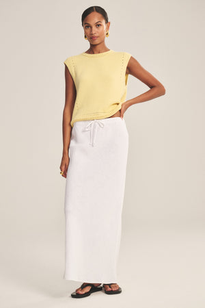 A woman wears the BEKA HEAVY LINEN SKIRT by Velvet by Graham & Spencer—a long white maxi skirt with a drawstring waist—paired with a sleeveless yellow knit top, black sandals, and gold hoop earrings.