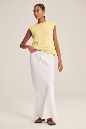 A woman stands against a plain background wearing a sleeveless yellow knit top, black sandals, gold hoop earrings, and the BEKA HEAVY LINEN SKIRT by Velvet by Graham & Spencer—a white maxi with a drawstring waist.