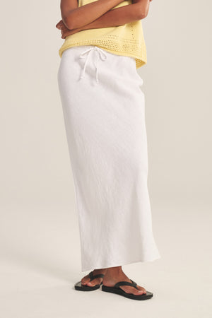 A person in a yellow sleeveless top, black sandals, and the BEKA HEAVY LINEN SKIRT by Velvet by Graham & Spencer—a white maxi with drawstring waist—stands with arms crossed.