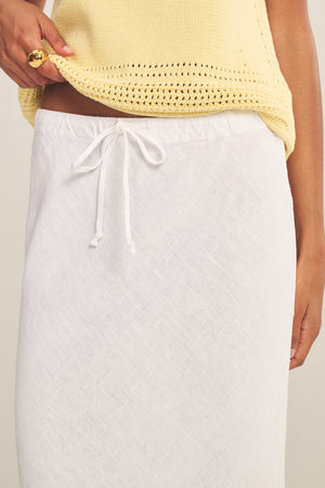 A person wearing a light yellow knit top and the BEKA HEAVY LINEN SKIRT by Velvet by Graham & Spencer, with one hand adjusting their shirt and the other resting at their side.