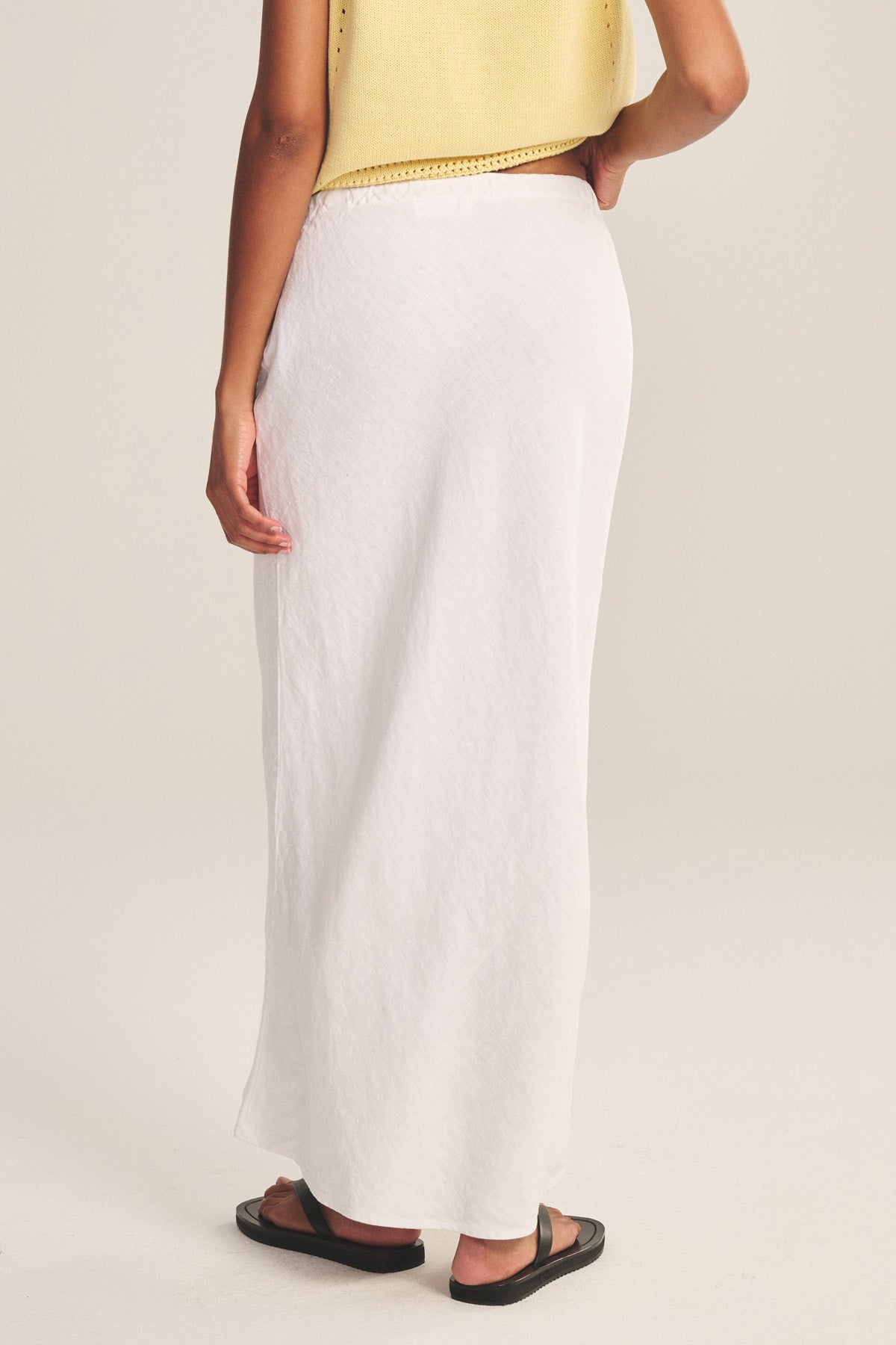 A person faces away, wearing a yellow sleeveless top, black sandals, and the BEKA HEAVY LINEN SKIRT by Velvet by Graham & Spencer—a maxi skirt with a drawstring waist—set against a plain background.-41128029913281