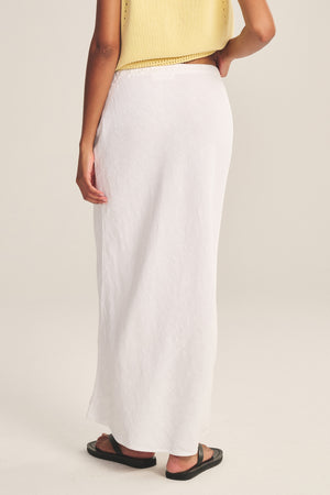 A person faces away, wearing a yellow sleeveless top, black sandals, and the BEKA HEAVY LINEN SKIRT by Velvet by Graham & Spencer—a maxi skirt with a drawstring waist—set against a plain background.