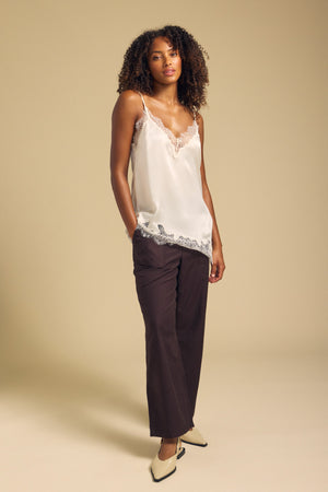 A woman stands against a beige background, wearing the BEVERLY SILK LACE CAMISOLE by Velvet by Graham & Spencer—with adjustable straps and lace trim—paired with dark brown pants and cream heels.