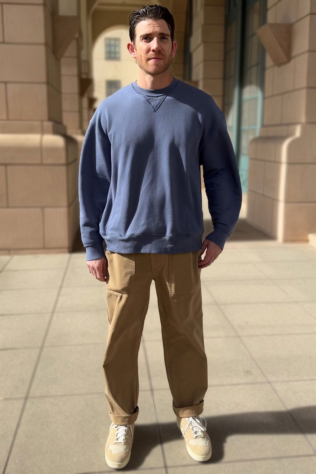 A man stands on a sunlit, tiled walkway wearing the FORREST SWEATSHIRT by Velvet by Graham & Spencer, paired with tan pants and beige sneakers, against a backdrop of stone walls and windows.-38326124282049