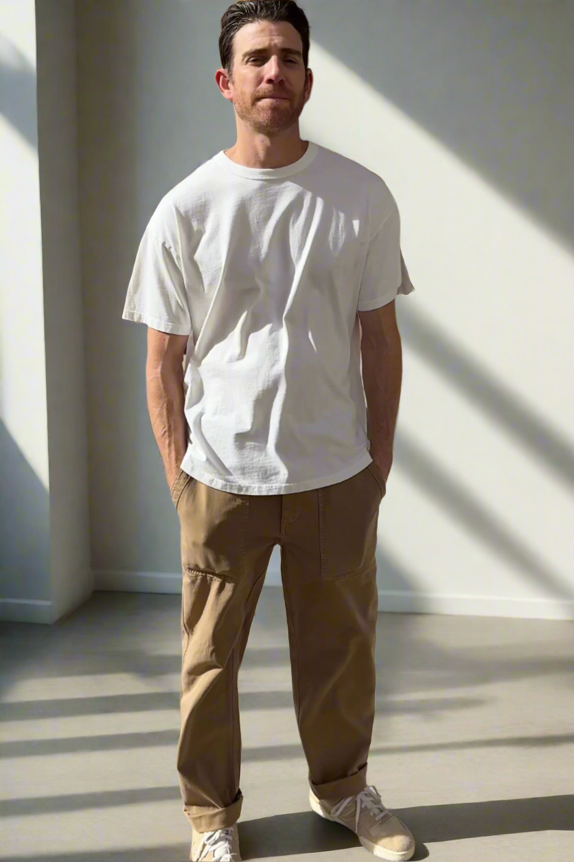   In a sunlit room, a man highlights everyday versatility by wearing the BRYAN TEE from Velvet by Graham & Spencer, crafted from premium pima cotton. He pairs it with brown pants and sneakers, displaying a relaxed fit that exudes casual ease as he comfortably tucks his hands in his pockets. 