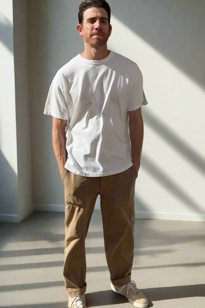 In a sunlit room, a man highlights everyday versatility by wearing the BRYAN TEE from Velvet by Graham & Spencer, crafted from premium pima cotton. He pairs it with brown pants and sneakers, displaying a relaxed fit that exudes casual ease as he comfortably tucks his hands in his pockets.