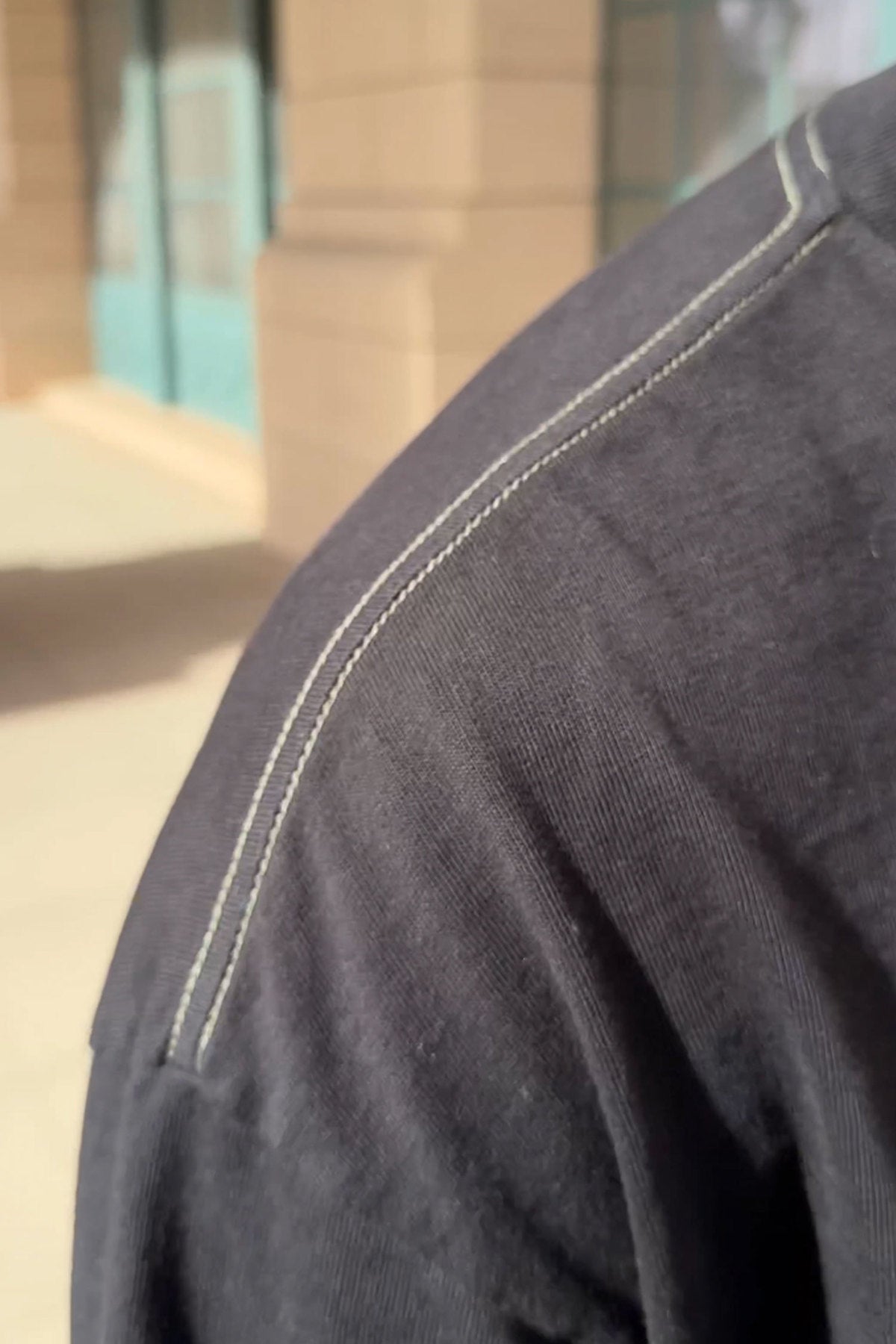   Close-up of a person wearing the Velvet by Graham & Spencer SLATER TEE in dark gray, showcasing its relaxed fit and visible shoulder seam stitching, while standing in a sunlit outdoor setting. 