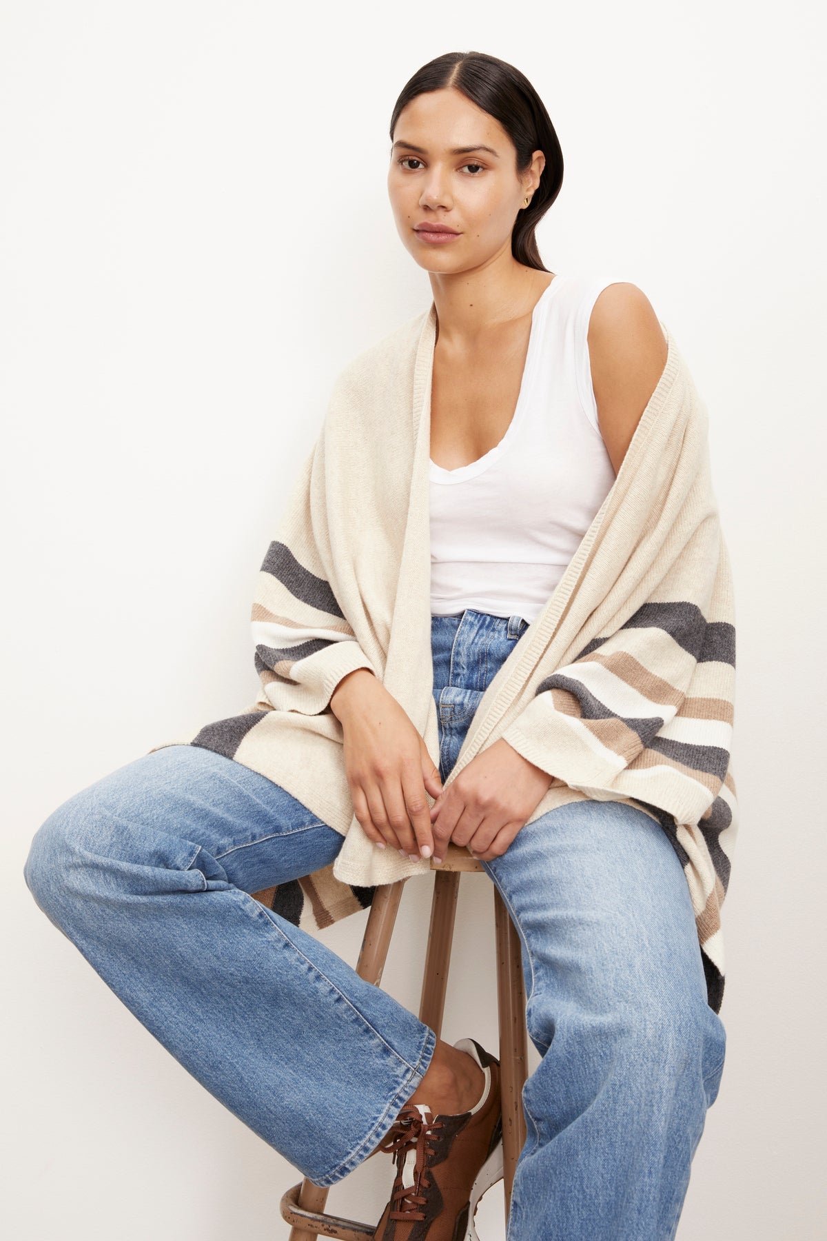 A woman is sitting on a stool wearing Velvet by Graham & Spencer's HARPER OPEN FRONT PONCHO and jeans, along with a striped cardigan.-26897792958657