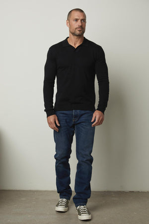 A man standing in a room wearing Velvet by Graham & Spencer jeans and a black v - neck sweater.