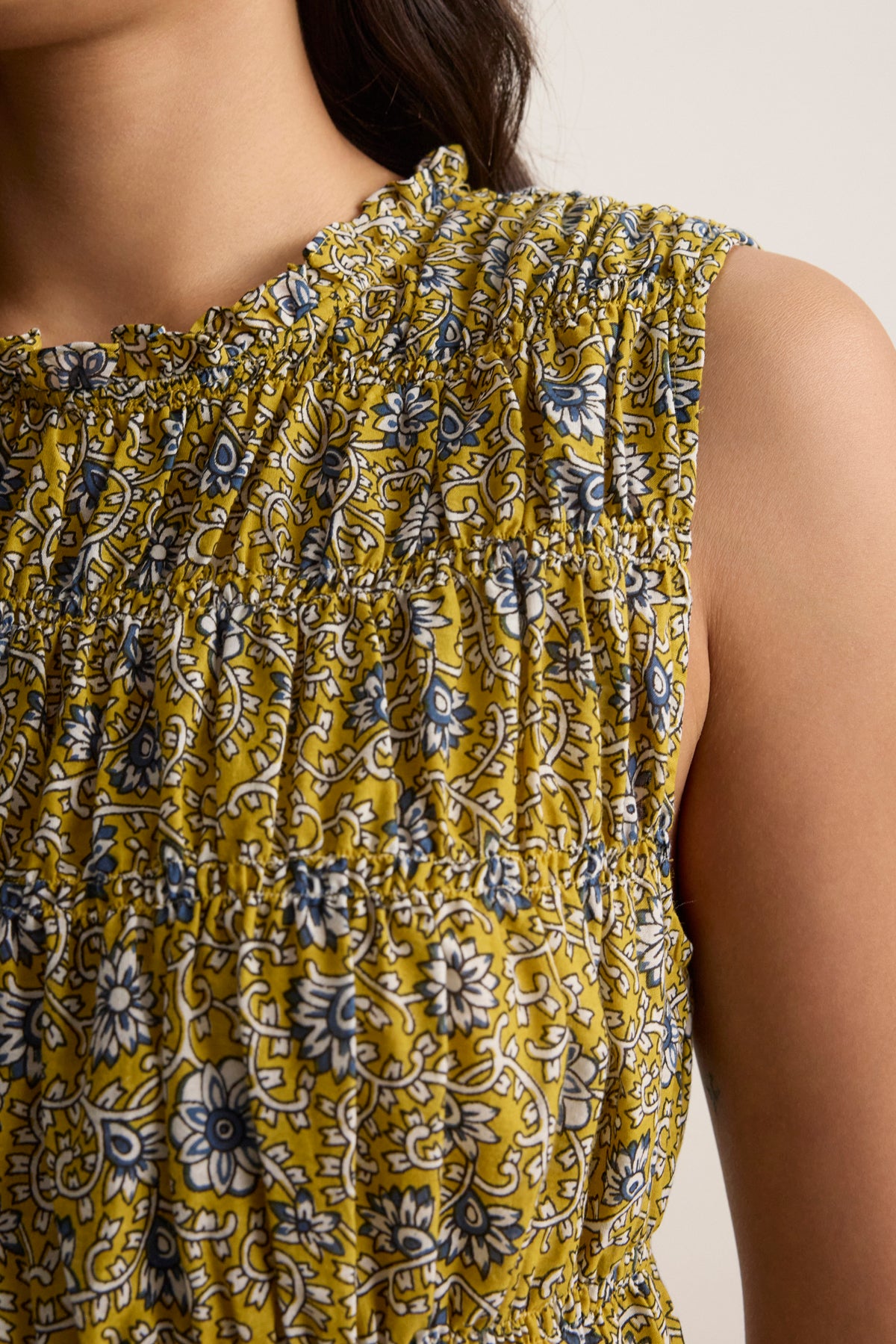 A close-up of a person wearing the MARLA DRESS by Velvet by Graham & Spencer—a sleeveless yellow mini dress with a blue and white floral pattern, block-printed cotton, and a ruffled round neckline.-39120095477953