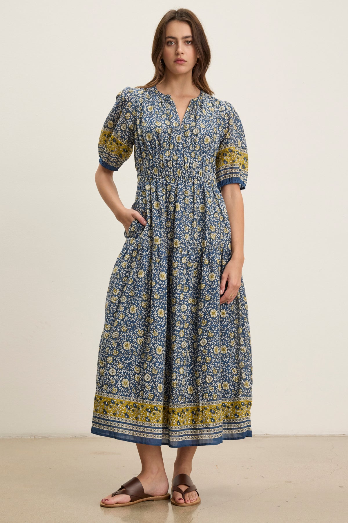 A woman poses against a plain background in the NATALIA DRESS by Velvet by Graham & Spencer—a blue and yellow floral cotton dress with a smocked waist, ankle-length hem, and short puff sleeves, styled with sandals.