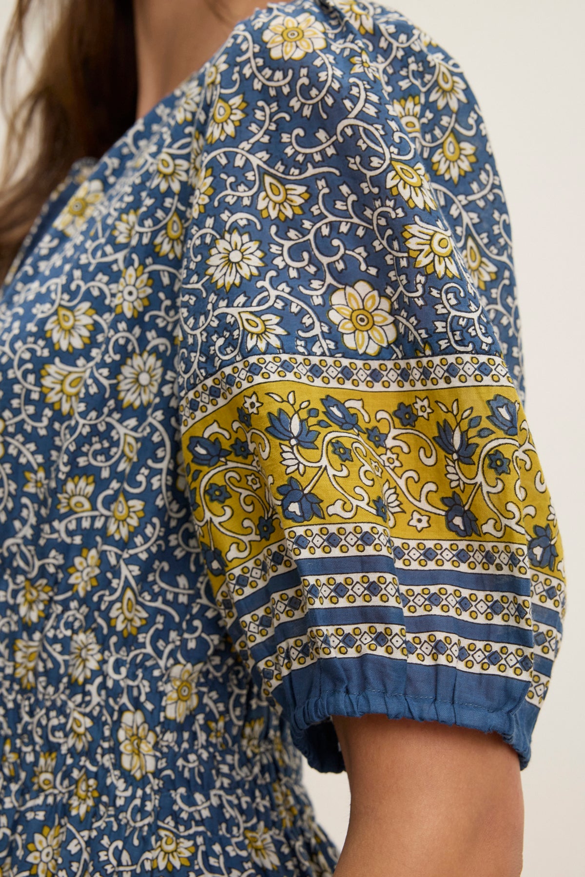 Close-up of a person wearing the NATALIA DRESS by Velvet by Graham & Spencer—a blue printed cotton dress with white and yellow florals, puffed short sleeves accented by a yellow band, and a smocked waist for shape.-Hover Image