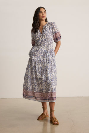 A woman stands indoors wearing the NATALIA DRESS by Velvet by Graham & Spencer, a printed cotton A-line with short sleeves and a v-neckline, styled with brown lace-up sandals.