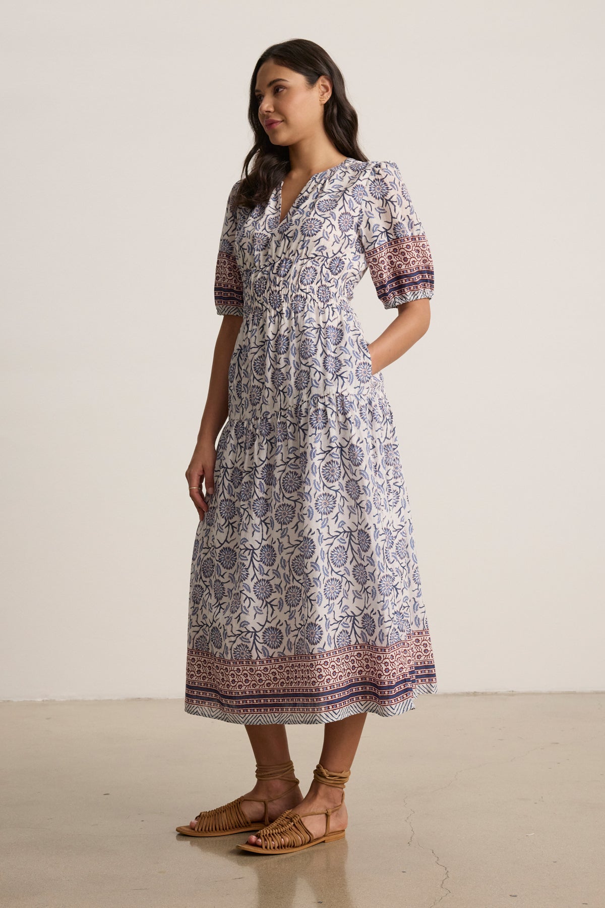   A woman wears the NATALIA DRESS by Velvet by Graham & Spencer—an ankle-length, patterned A-line piece with short puff sleeves—paired with sandals, standing on a light floor before a neutral background. 
