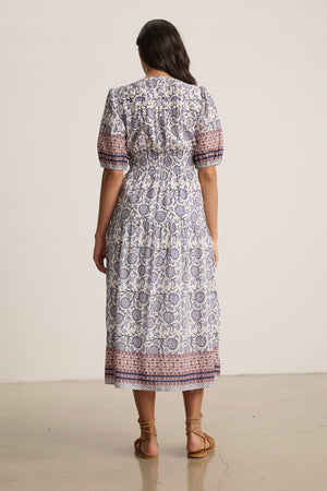 A woman faces away, wearing the NATALIA DRESS by Velvet by Graham & Spencer—a mid-length printed cotton dress with short sleeves—paired with strappy sandals against a plain, neutral background.