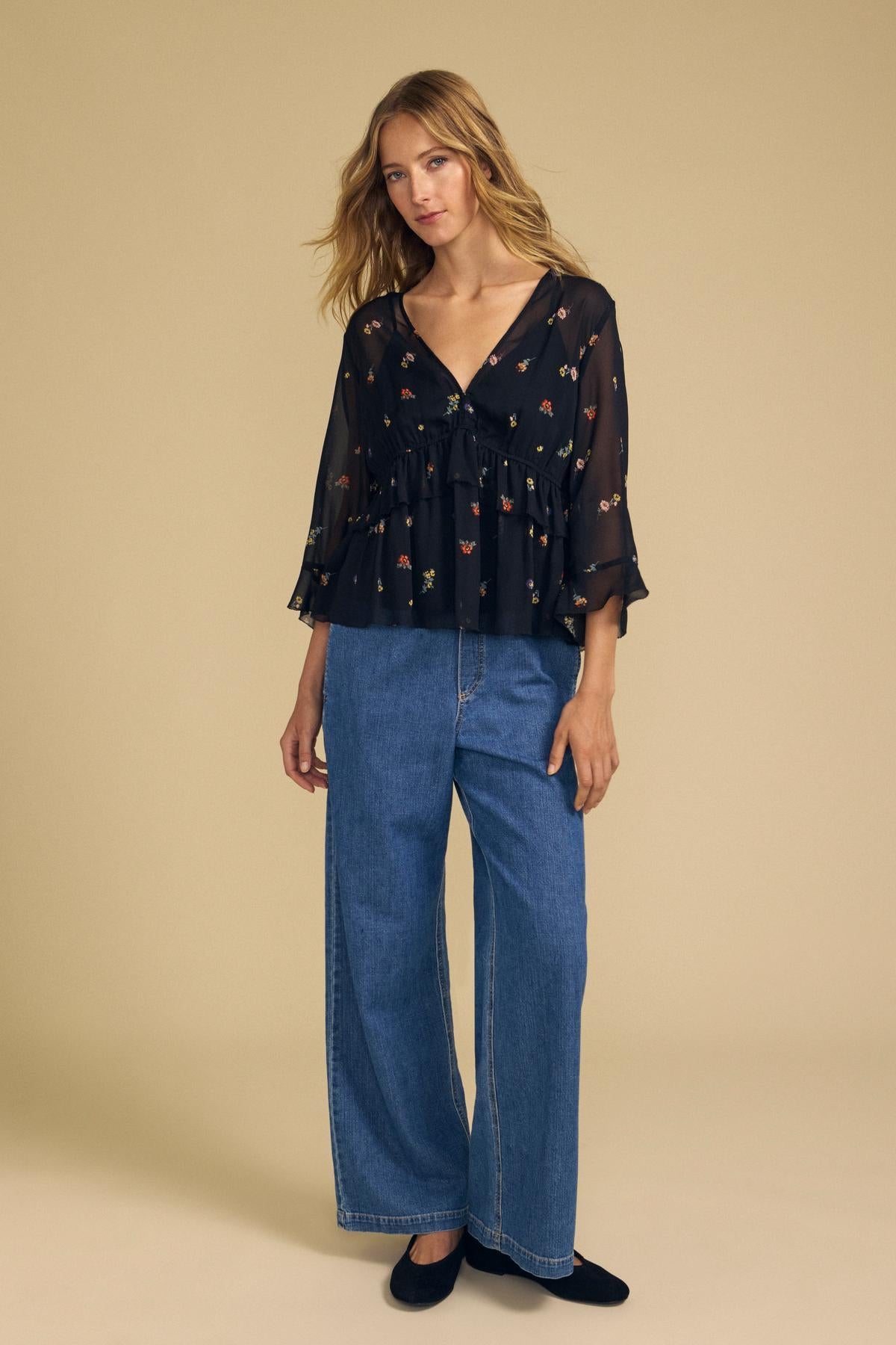 A woman stands against a beige background wearing the AILEEN BLOUSE by Velvet by Graham & Spencer, styled with wide-leg blue jeans and black shoes.
