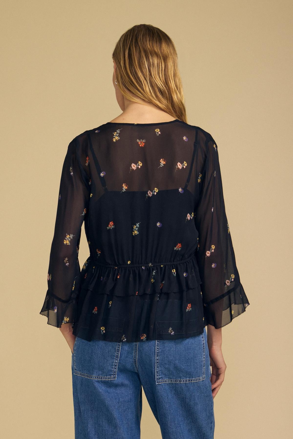 A person with long blonde hair is seen from the back against a beige background, wearing Velvet by Graham & Spencer’s AILEEN BLOUSE—a sheer viscose floral printed blouse with ruffled sleeves—paired with blue jeans.
