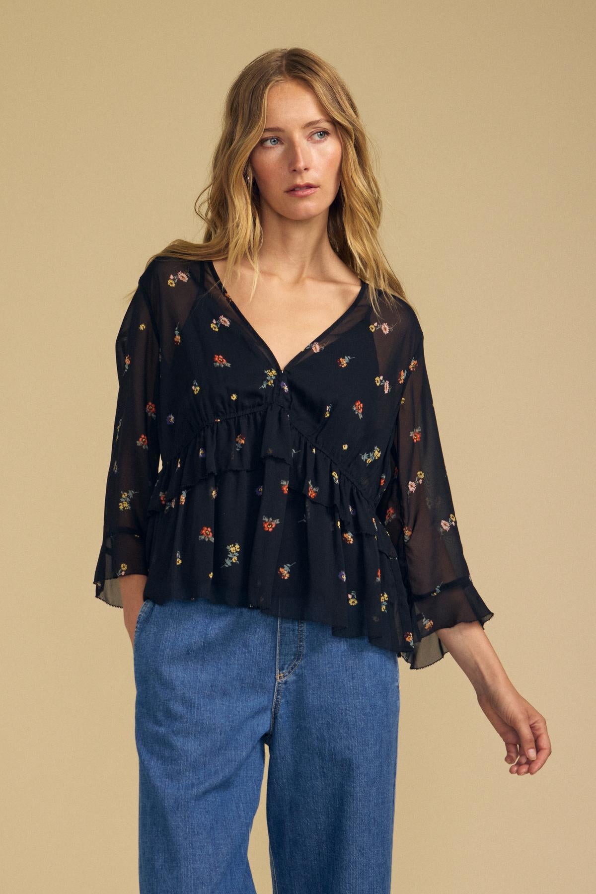 A woman wearing the AILEEN BLOUSE by Velvet by Graham & Spencer, featuring sheer viscose fabric with a floral print, ruffled details, and wide sleeves, paired with blue jeans and standing against a beige background.