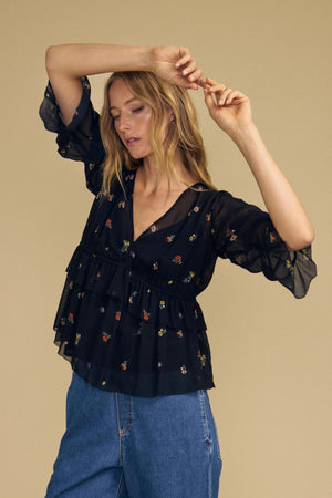 Model wearing the AILEEN BLOUSE by Velvet by Graham & Spencer—a sheer viscose top featuring black floral prints and ruffle details—paired with blue jeans, poses with arms raised against a beige background.