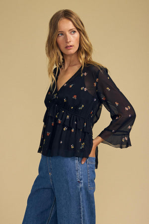A woman stands against a beige background, wearing the Velvet by Graham & Spencer AILEEN BLOUSE—a black, floral-printed, long-sleeve top—paired with blue jeans. Her left hand is in her pocket as she gazes slightly to the side.