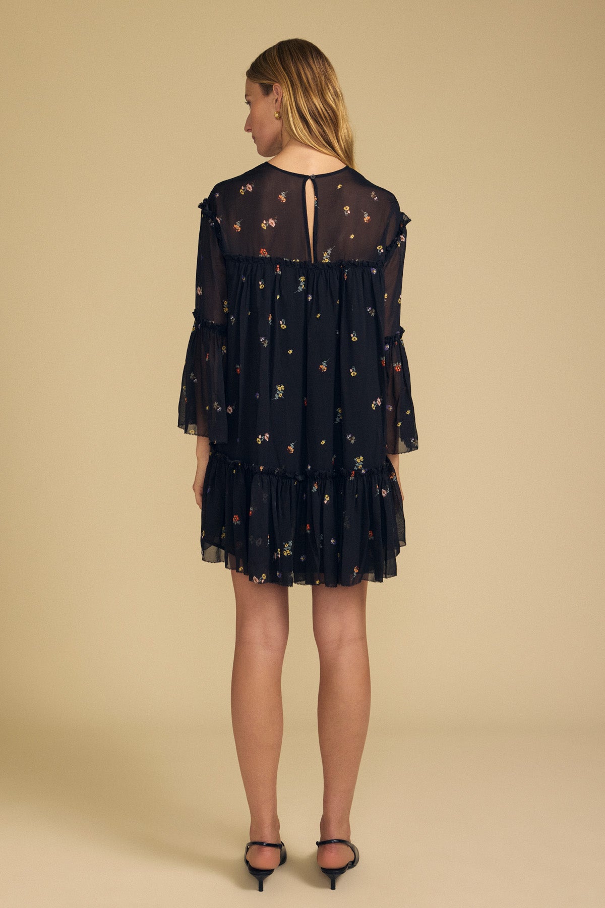 A woman stands facing away, wearing the CLARICE DRESS by Velvet by Graham & Spencer—a lightweight summer dress featuring sheer sleeves and a delicate small floral pattern—paired with black sandals, set against a plain beige background.-41946779615425