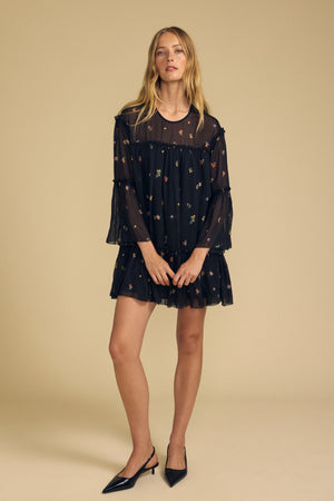 A woman stands in a studio against a beige background, wearing the CLARICE DRESS by Velvet by Graham & Spencer—a black, sheer viscose dress with long sleeves and a knee-length hem. Delicate floral embroidery details and a lined, boxy silhouette lend the look modern elegance. She completes the outfit with black pointed heels.