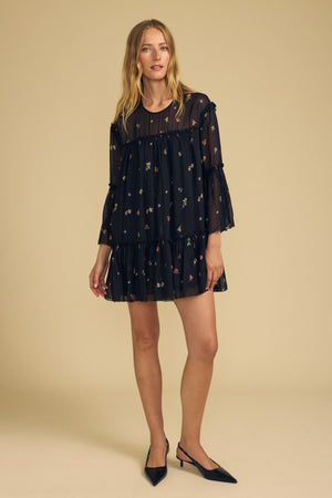 A woman wearing the CLARICE DRESS by Velvet by Graham & Spencer—a black sheer viscose dress adorned with small floral patterns and ruffled tiers—paired with black pointed-toe flats, stands against a plain beige background.