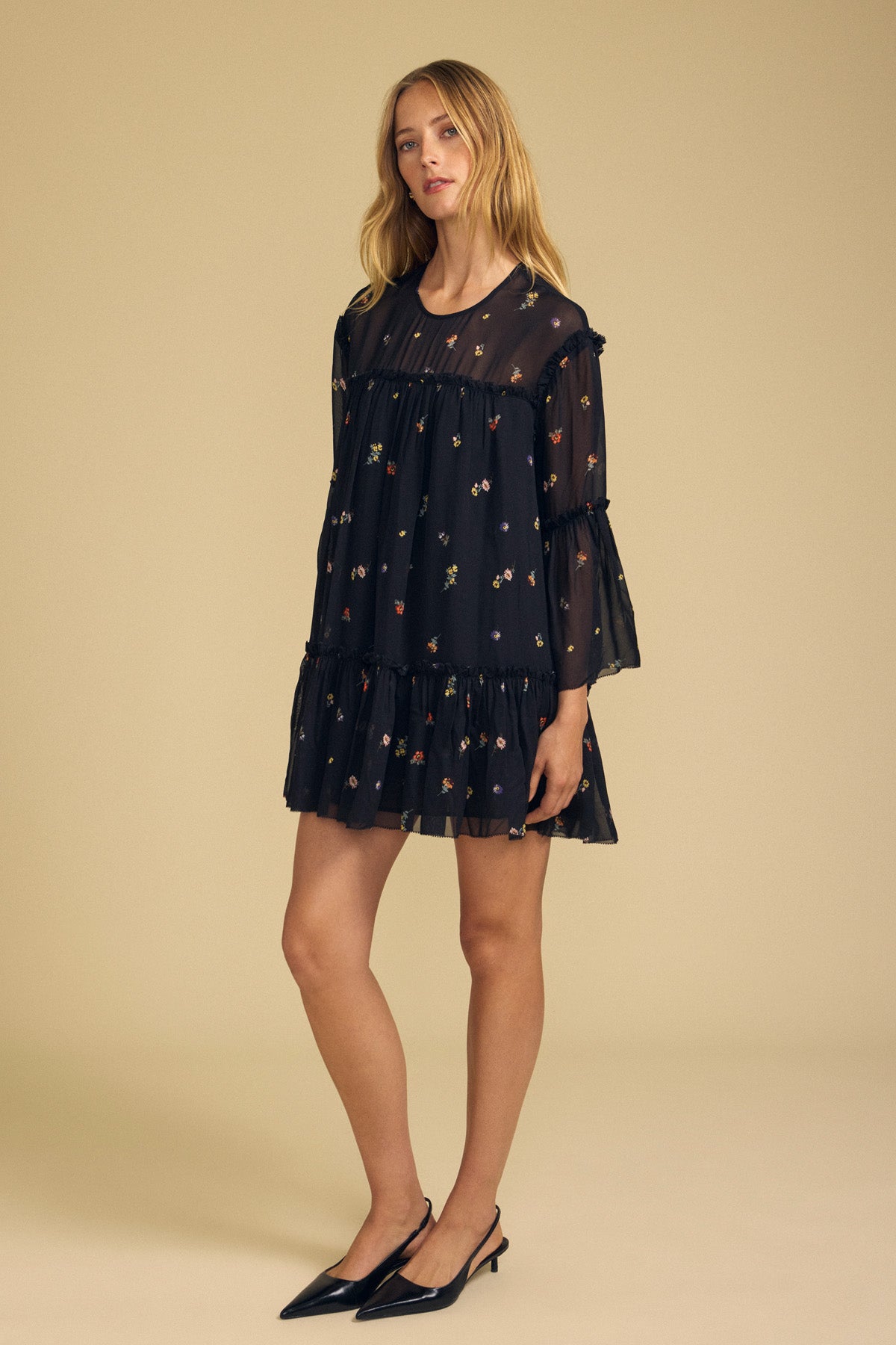 A woman stands in a studio wearing the CLARICE DRESS by Velvet by Graham & Spencer—a loose, sheer viscose dress featuring delicate floral prints and ruffled layers—paired with black pointed heels.-41946779418817