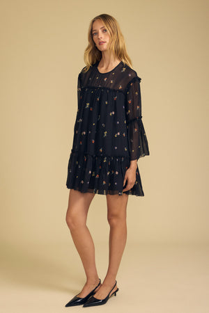 A woman stands in a studio wearing the CLARICE DRESS by Velvet by Graham & Spencer—a loose, sheer viscose dress featuring delicate floral prints and ruffled layers—paired with black pointed heels.
