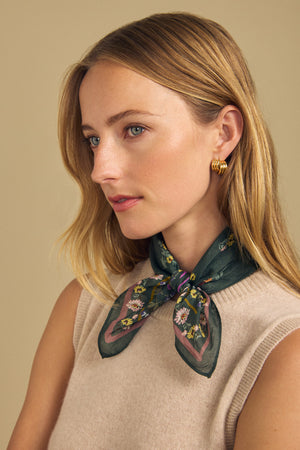 A woman with blonde hair wearing a sleeveless beige top and the MARIANNA SCARF by Velvet by Graham & Spencer—a green floral printed scarf—tied at her neck, along with gold hoop earrings, looks to the side.