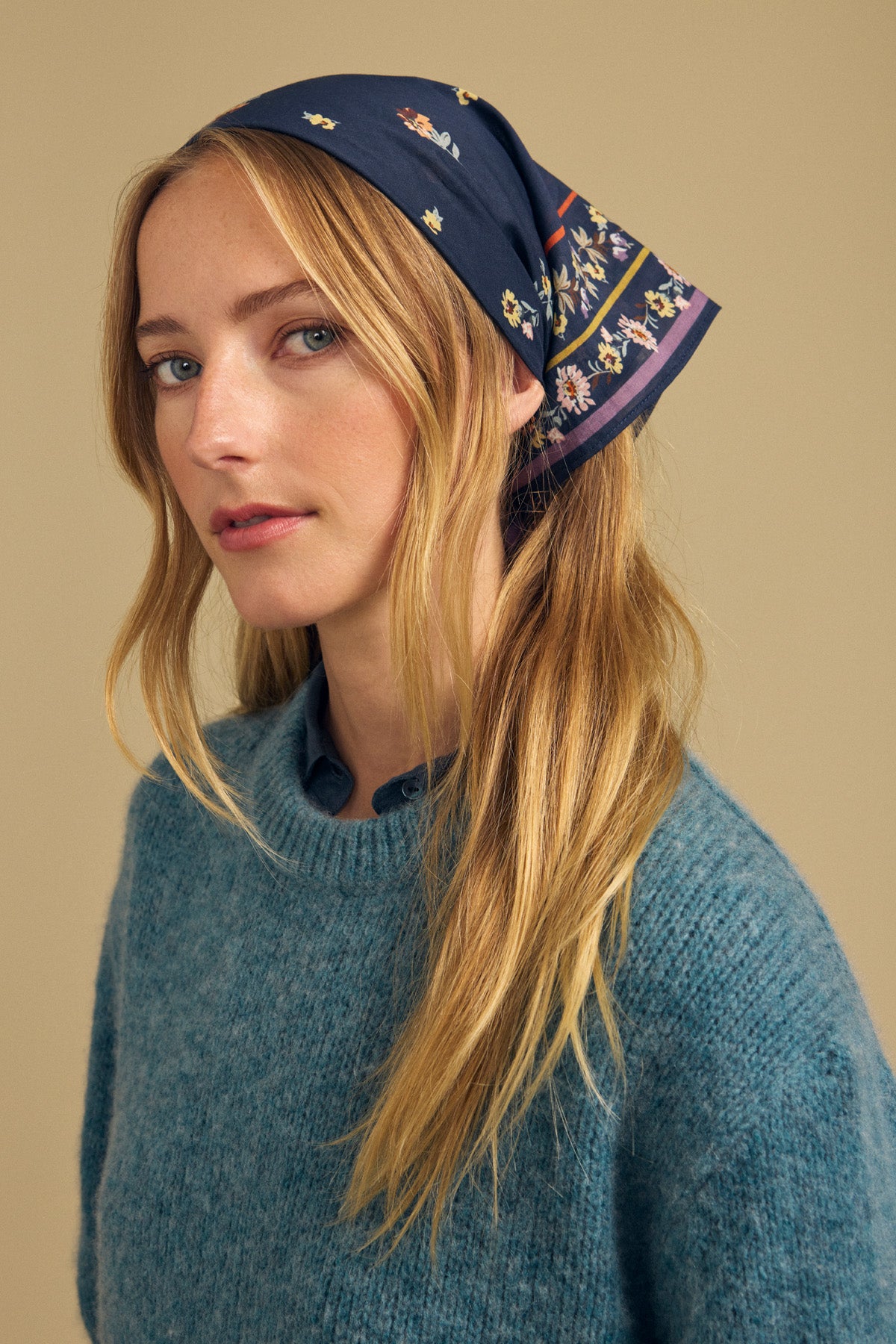A woman with long blonde hair wears a blue sweater and the MARIANNA SCARF by Velvet by Graham & Spencer—a lightweight, navy patterned scarf styled as a head wrap—posing against a plain beige background.