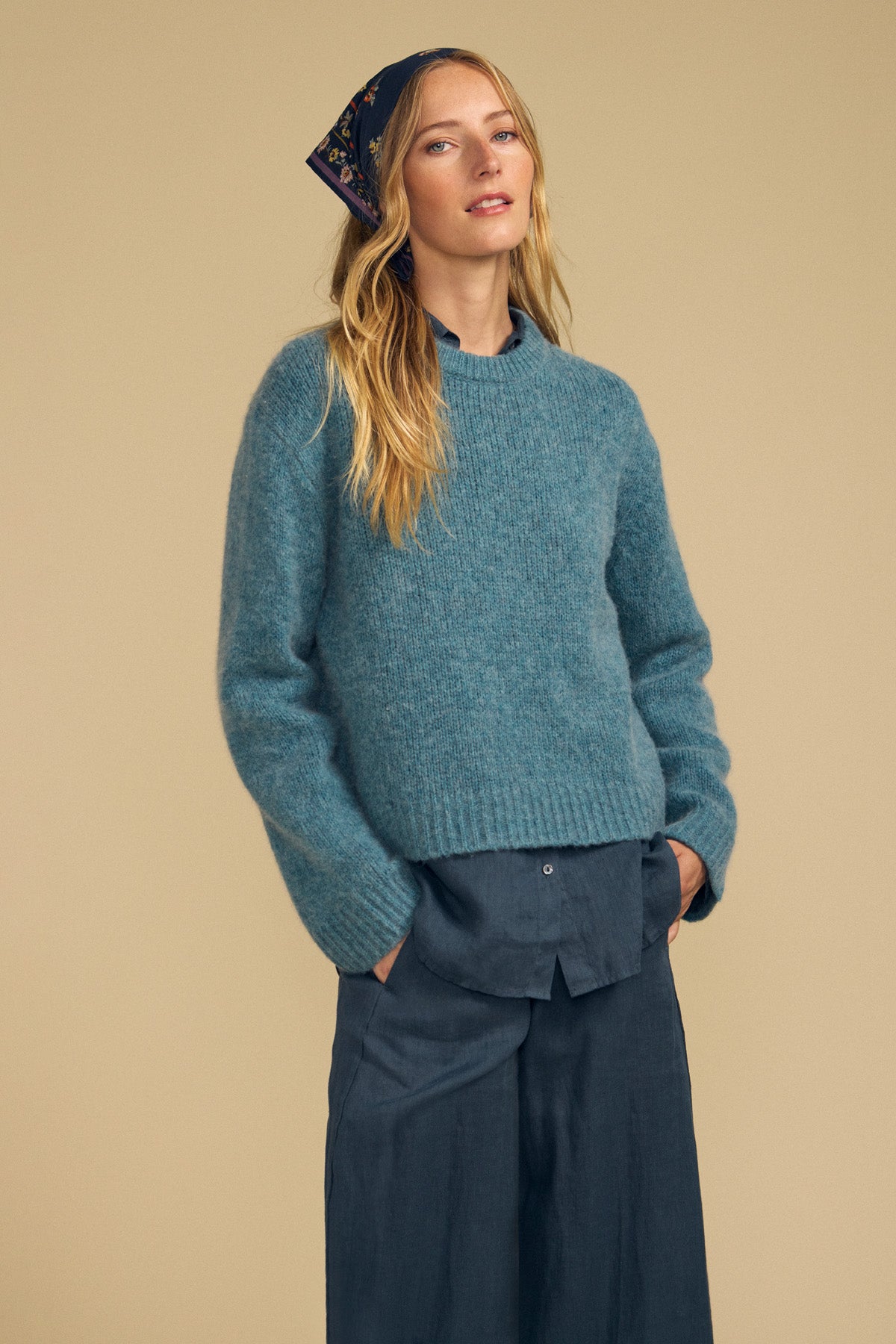 A person with long blonde hair stands against a plain beige background with hands in their pockets, wearing a blue sweater layered over a collared shirt, wide-leg pants, and the MARIANNA SCARF by Velvet by Graham & Spencer styled as a floral printed headscarf.-41964669370561