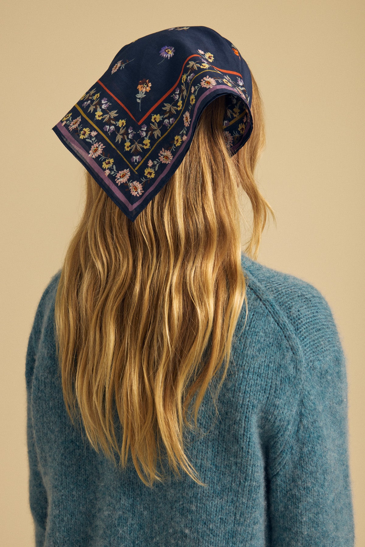 A person with long, wavy blonde hair, facing away from the camera, wears the Velvet by Graham & Spencer MARIANNA SCARF—a dark blue floral square scarf—as a head accessory along with a blue knit sweater.