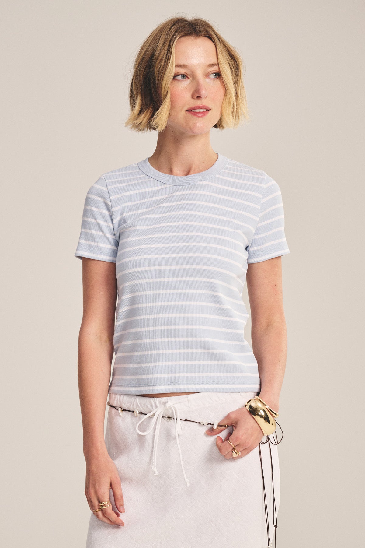 A woman wears the BRENDA TEE by Velvet by Graham & Spencer, a light blue and white striped cropped fitted top with a ribbed crew neckline, paired with a drawstring white skirt against a plain backdrop.