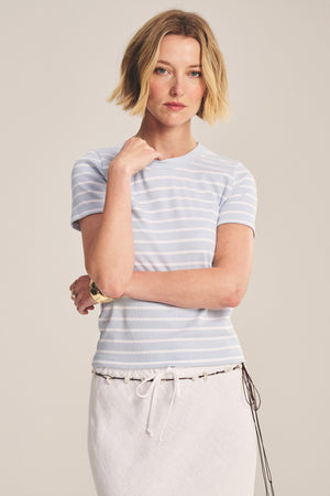 A woman with short blonde hair wears the BRENDA TEE by Velvet by Graham & Spencer—a light blue and white striped cropped top with a ribbed crew neckline—paired with a white skirt, standing with one hand touching her face.