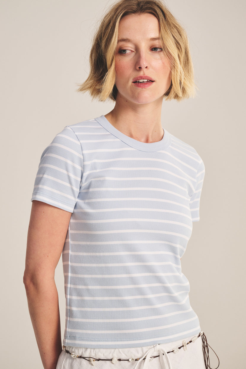 A woman with short blonde hair wears the BRENDA TEE by Velvet by Graham & Spencer—a light blue and white striped cropped tee with a ribbed crew neckline—paired with light-colored pants against a neutral background.