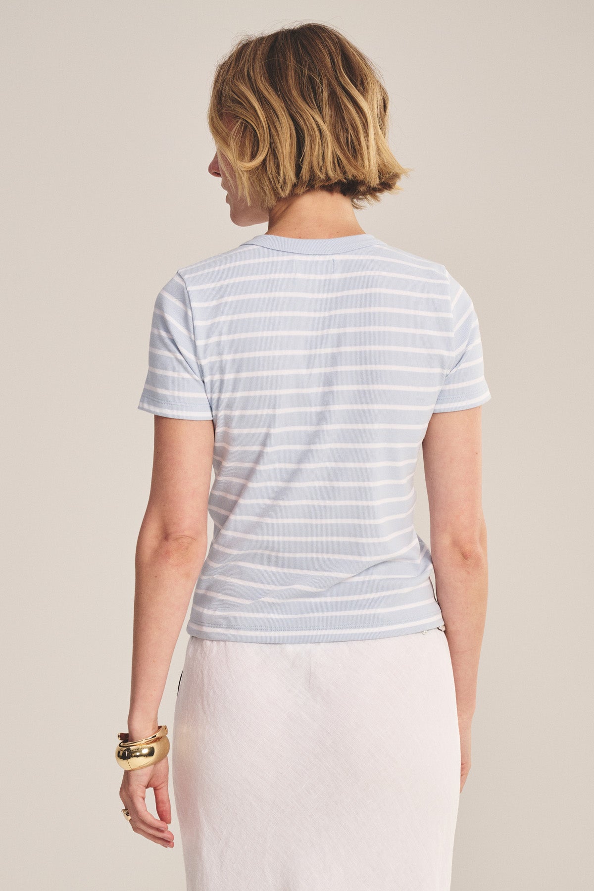 A woman with short, light brown hair is shown from the back wearing the BRENDA TEE by Velvet by Graham & Spencer—a light blue and white striped, fitted cropped tee—paired with a white skirt against a neutral background.-41338350764225