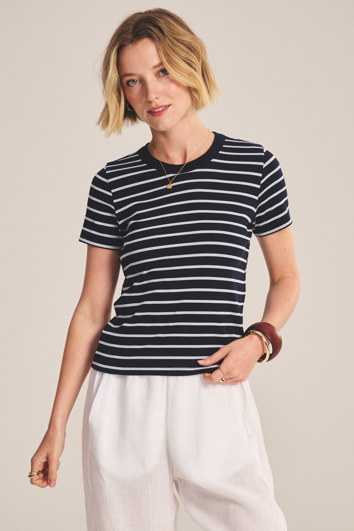 Wearing the BRENDA TEE by Velvet by Graham & Spencer, a black and white striped cropped fitted top, paired with white pants, the model stands against a plain background.