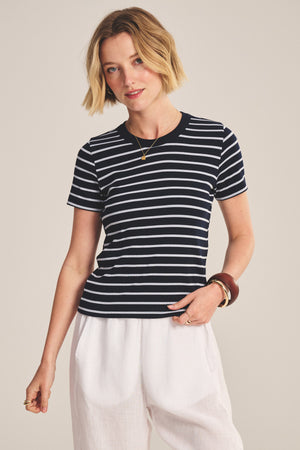 Wearing the BRENDA TEE by Velvet by Graham & Spencer, a black and white striped cropped fitted top, paired with white pants, the model stands against a plain background.