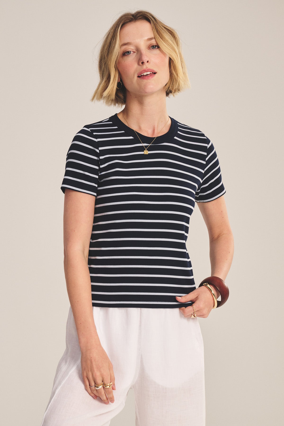 A person with short blonde hair wears the BRENDA TEE by Velvet by Graham & Spencer, a black and white striped cropped tee with a fitted silhouette, paired with white pants and jewelry, standing against a plain background.