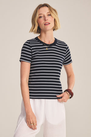 A person with short blonde hair wears the BRENDA TEE by Velvet by Graham & Spencer, a black and white striped cropped tee with a fitted silhouette, paired with white pants and jewelry, standing against a plain background.