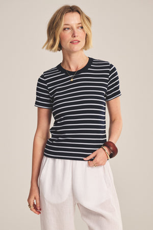 A person wears the BRENDA TEE by Velvet by Graham & Spencer—a black and white striped, cotton rib cropped top—paired with white pants against a neutral background.