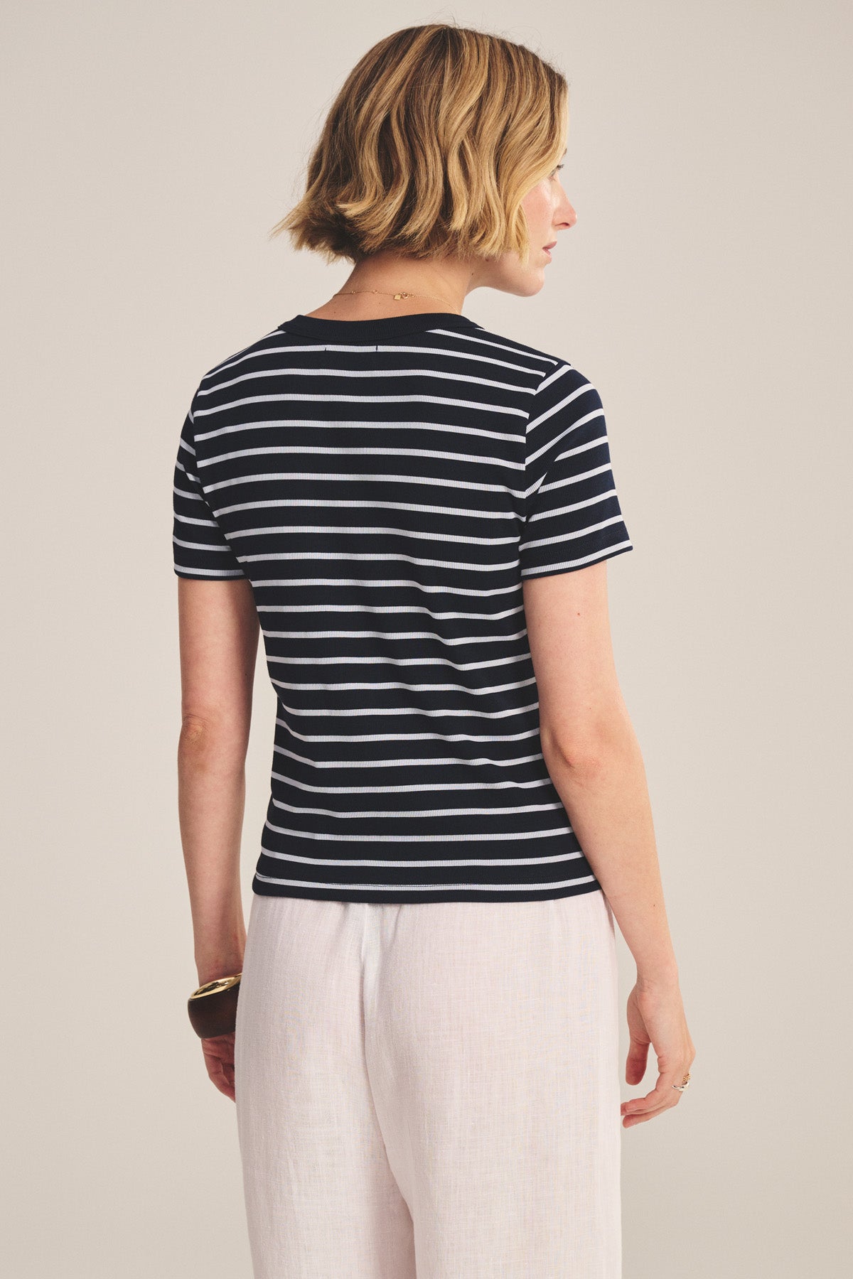 A woman with short blonde hair, wearing the BRENDA TEE by Velvet by Graham & Spencer and light-colored pants, stands facing away from the camera.-40959850447041
