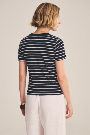 A woman with short blonde hair, wearing the BRENDA TEE by Velvet by Graham & Spencer and light-colored pants, stands facing away from the camera.