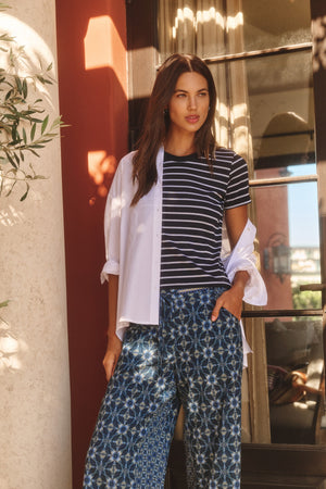 Outdoors, a woman wears the Velvet by Graham & Spencer BRENDA TEE—a navy striped cropped top with a fitted silhouette—layered over a white button-down shirt and paired with blue patterned wide-leg pants, one hand in her pocket.